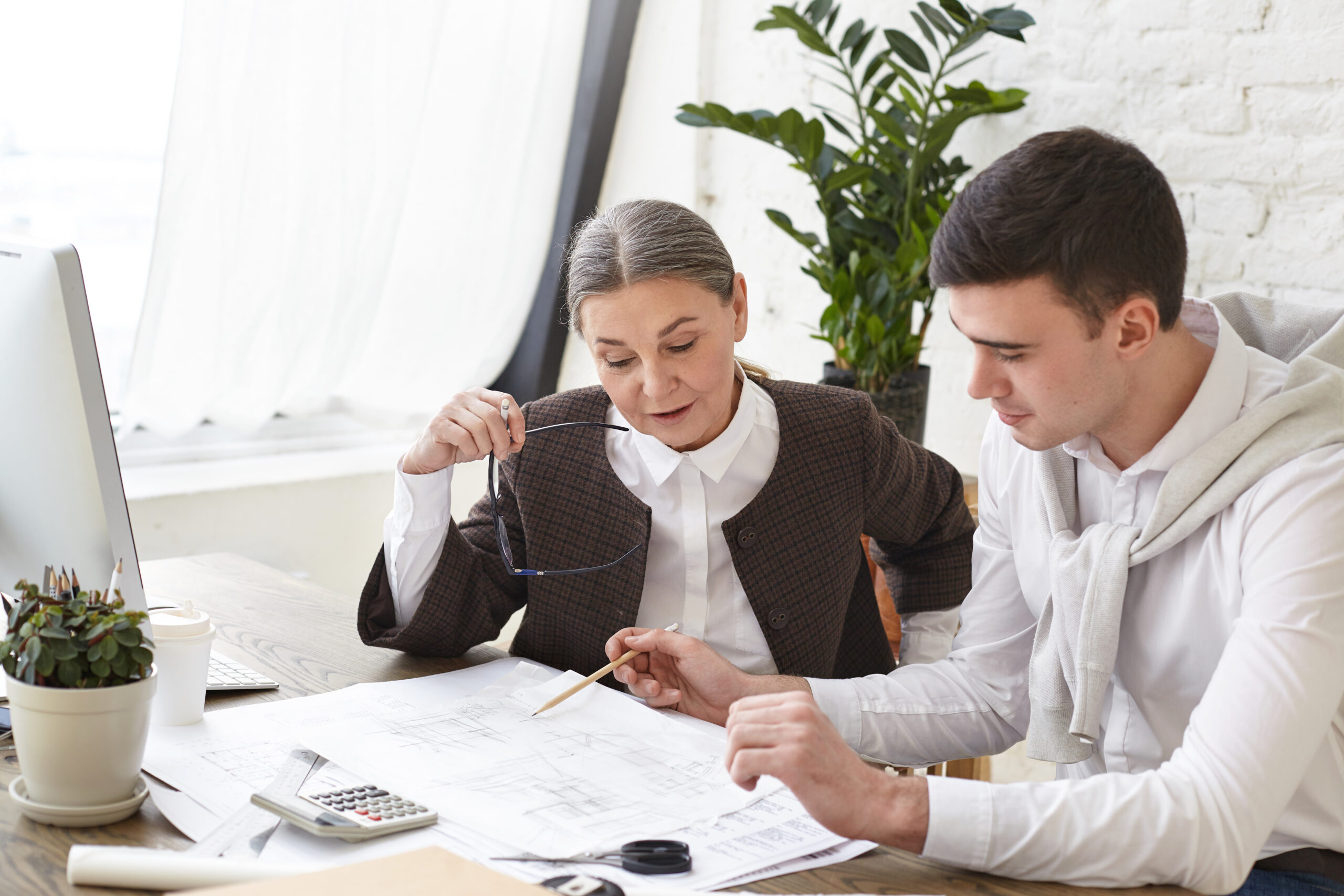 Architecture, construction and engineering. Candid shot of mature woman with grey hair sitting at her workplace and listening to young male architect, who explaining something in his drawings Architecture, construction and engineering. Candid shot of mature woman with grey hair sitting at her workplace and listening to young male architect, who explaining something in his drawings